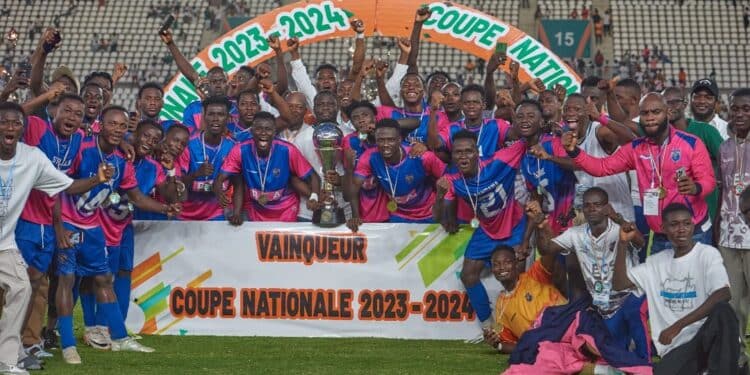 Image de Football. La 58e édition de la Coupe nationale de Côte d’Ivoire a couronné le Racing Club d’Abidjan, qui a triomphé sur l’ASEC Mimosas au Stade de la Paix de Bouaké. Cette victoire historique a été scellée par un but crucial de Bandama Yao Junior, marqué sur penalty à la 73e minute, offrant au club de la capitale son premier titre national. Une Victoire Historique Le Racing Club d’Abidjan, longtemps considéré comme un challenger potentiel dans le paysage du football ivoirien, a marqué l’histoire en remportant pour la première fois la Coupe nationale. Le match décisif contre l’ASEC Mimosas, réputé pour sa domination historique dans le football ivoirien, s’est déroulé sous une tension palpable au Stade de la Paix de Bouaké. Bandama Yao Junior, l’auteur du seul but de la rencontre, a été l’artisan principal de cette victoire mémorable. « Nous avons travaillé dur pour en arriver là, et cette victoire est le fruit de notre détermination et de notre cohésion d’équipe », a déclaré le capitaine du Racing Club dans une interview après le match. L’ASEC Mimosas, malgré cette défaite amère, se console avec une qualification pour la prochaine Coupe de la Confédération. Sol Béni, entraîneur emblématique de l’équipe, a exprimé à la presse sa déception et sa fierté : « Bien que nous ayons échoué dans cette finale, nous tournons notre regard vers l’avenir avec l’objectif de rebondir et de faire honneur à nos supporters dans les compétitions à venir. » Perspectives pour le Football Ivoirien La victoire du Racing Club d’Abidjan soulève des questions sur l’évolution du paysage footballistique en Côte d’Ivoire. Cette performance pourrait marquer le début d’une nouvelle ère de compétitivité accrue entre les clubs traditionnels et les équipes émergentes. Comment cette victoire influencera-t-elle les stratégies de recrutement et les ambitions des clubs pour les saisons futures ? La 58e édition de la Coupe nationale restera gravée dans l’histoire du football ivoirien grâce à la performance exceptionnelle du Racing Club d’Abidjan. Alors que les supporters célèbrent ce succès tant attendu, le football du pays entre dans une période de réflexion et de réajustement stratégique. Quelles leçons les autres clubs peuvent-ils tirer de cette victoire ?