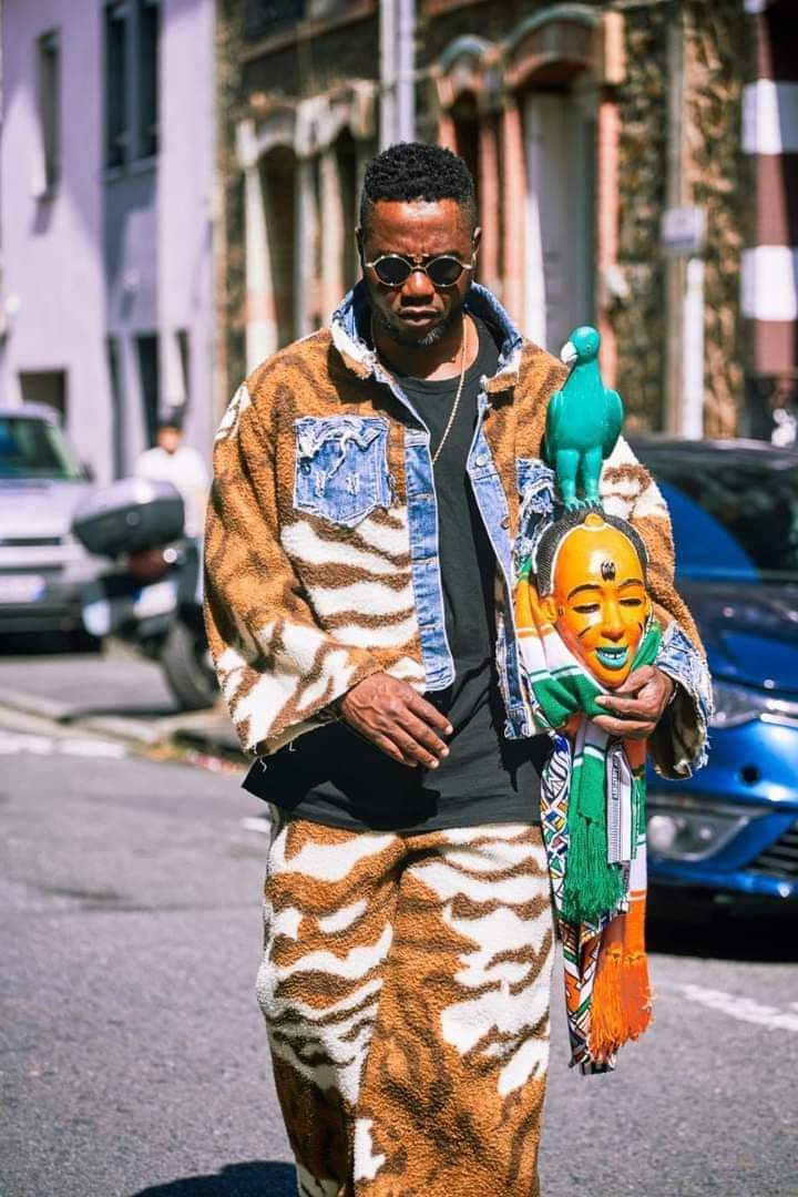 Image de Célébrités. L'artiste danseur chorégraphe ivoirien, Gadoukou La Star a été aperçu dans les rues de Vienne, en Autriche, avec un masque ivoirien en main et dans une tenue qui rappelle l'Afrique. C'est peut être un moyen pour lui d'afficher son africanité sur cette terre occidentale. Au moins, il ne renie pas sa culture mais au contraire l'affiche publiquement. Que pensez-vous de ceux qui, vivant en Europe, s'expriment et s'habillent comme des européens ?